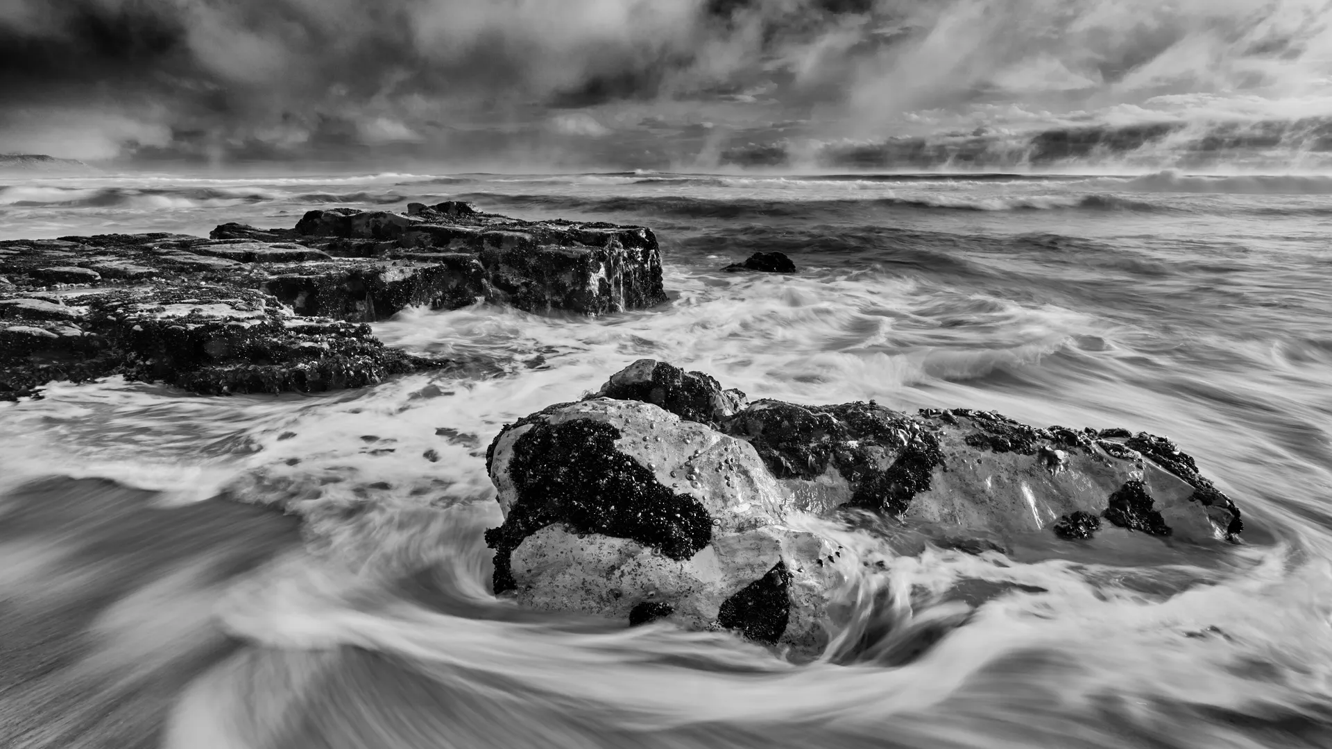 Black and white long exposure of waves swirling around rocks under stormy skies, Misty Cliff, Cape Region, South Africa